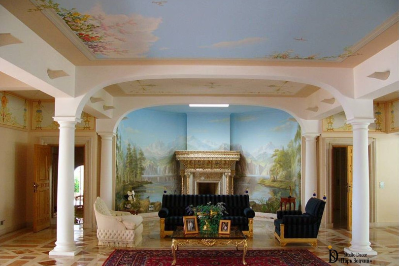 Роспись полностью изменила представления о размерах помещения, стены и потолки потеряли свои очертания, сделали тем самым зону отдыха яркой и «свободной» от внешних ограничений Интерьер гостиной с камином, колоннами и росписью